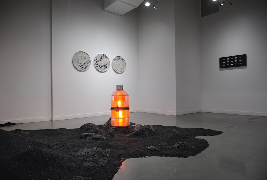 Glowing orange cylindrical light sculpture displayed in a minimalist gallery space with framed artworks on the walls.
